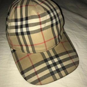 Rutig keps från Burberry - Snygg rutig keps från Burberry i klassiskt beige, svart och rött mönster. Kepsen har en justerbar rem baktill för perfekt passform. Perfekt för både vardag och festliga tillfällen. Ett måste för den stilmedvetna!