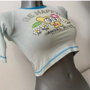 Blå croppad topp från Angel Blue - Supergullig ljusblå croppad topp från Angel Blue med ett färgglatt tryck av en söt figur och texten 'BE HAPPY!'. Perfekt för en avslappnad och glad stil. Den har korta ärmar och blå detaljer vid hals och ärmslut🌼