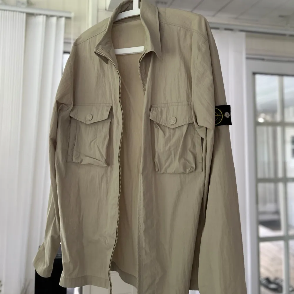 Säljer en snygg beige jacka från Stone Island. Den har en dragkedja framtill och två praktiska bröstfickor med knappar. Jackan är långärmad och har den klassiska Stone Island-loggan på ärmen. Perfekt för vår och höst! Diskuterar priset🤍. Jackor.