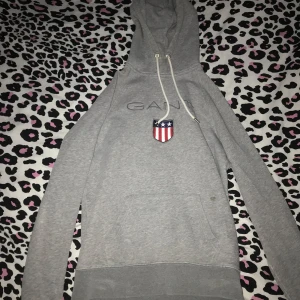 Grå hoodie från GANT - Säljer en snygg och bekväm grå hoodie från GANT. Den har en klassisk design med en känguruficka och ett coolt emblem på bröstet. Perfekt för en avslappnad stil!