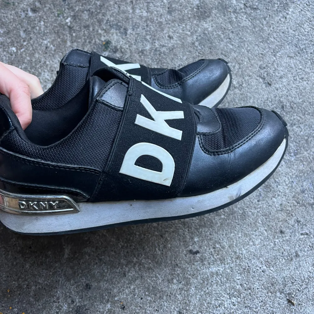 Säljer ett par snygga svarta sneakers från DKNY. De har en cool design med stora vita bokstäver på ovansidan och en bekväm passform. Skorna är i syntetmaterial och har en vit sula som ger en skön kontrast. Perfekta för både vardag och fest!. Skor.