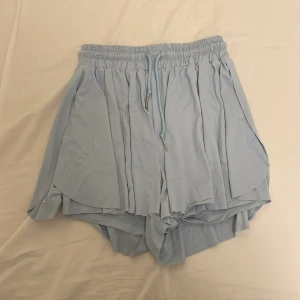 Ljusblå shorts med knytband - Säljer ett par superbekväma ljusblå shorts med elastisk midja och knytband. De har en lös passform och är perfekta för sommardagar. Materialet känns lätt och luftigt, vilket gör dem idealiska för både vardag och semester.