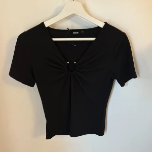 Svart topp från Bershka - Snygg svart topp från Bikbok med en v-ringad design och en cool detalj framtill. Toppen har korta ärmar och en tight passform, perfekt för både vardag och fest. Materialet känns mjukt och stretchigt.