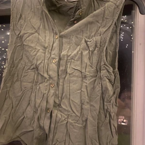 Grön ärmlös skjorta från H&M - Säljer en grön ärmlös skjorta från H&M. Skjortan har knappar framtill och en bröstficka. Perfekt för varma dagar eller som ett lager under en jacka. Den är i ett välanvänt skick men fortfarande snygg och användbar.