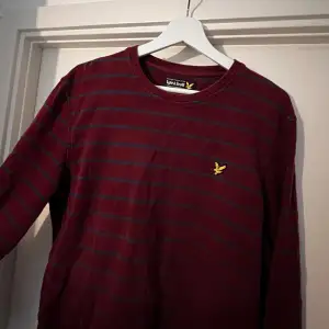 Säljer en snygg vinröd tröja från Lyle & Scott med mörkblå ränder. Den har en klassisk rund hals och ett gult logomärke på bröstet. Perfekt för både vardag och lite finare tillfällen. Tröjan är i storlek L och är superbekväm att ha på sig. 