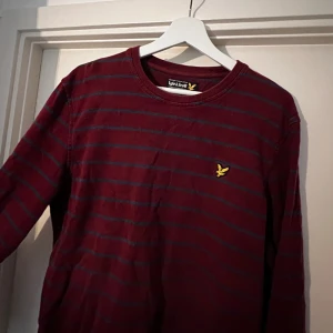 Vinröd randig tröja från Lyle & Scott - Säljer en snygg vinröd tröja från Lyle & Scott med mörkblå ränder. Den har en klassisk rund hals och ett gult logomärke på bröstet. Perfekt för både vardag och lite finare tillfällen. Tröjan är i storlek L och är superbekväm att ha på sig. 