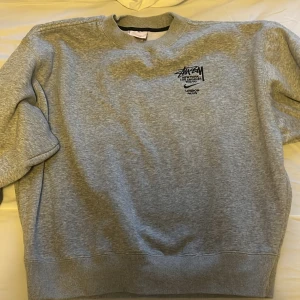 Grå sweatshirt från Nike x Stüssy - Säljer en grå sweatshirt från Nike i samarbete med Stüssy. Tröjan har en klassisk rund halsringning och är dekorerad med Stüssys ikoniska logga och städer som New York, Los Angeles, Tokyo, London och Paris. Vid snabb affär 550kr