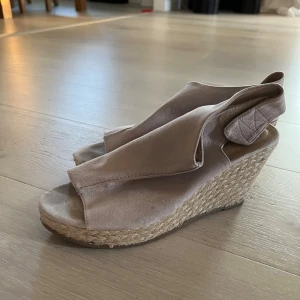 Beige kilklack sandaler - Säljer ett par snygga beige sandaler med kilklack. De har en öppen tå och är gjorda i ett mjukt material som ser ut som mocka. Perfekta för sommaren och passar både till vardags och fest. De har en bekväm passform och en stilren design.