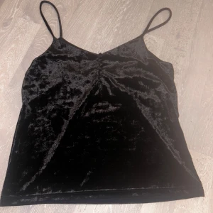 Svart sammetstopp från Lindex - Säljer en elegant svart sammetstopp från Lindex. Den är ärmlös med smala axelband och har en snygg, glansig finish. Perfekt för festliga tillfällen eller en utekväll. Toppen är i storlek XS och i nyskick.