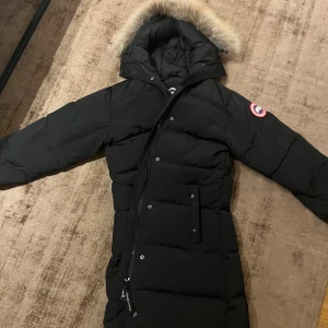Svart dunjacka från Canada Goose - Säljer en svart dunjacka från Canada Goose med en mysig pälskantad huva. Jackan är perfekt för kalla vinterdagar och har en snygg, lång design med dragkedja och knappar. Den har också en ikonisk logga på ärmen. Supervarm och stilren!