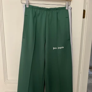 Gröna byxor från Palm Angels - Säljer ett par gröna byxor från Palm Angels. De har en avslappnad passform med elastisk midja och vita sidopaneler. Byxorna är gjorda av polyester och är märkta som eco-friendly.