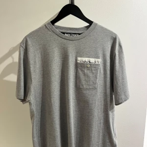 Grå t-shirt från Palm Angels - Säljer en helt ny stilren grå t-shirt från Palm Angels. Den har en liten bröstficka med en knappdetalj och märkeslogga. Perfekt för en avslappnad look!