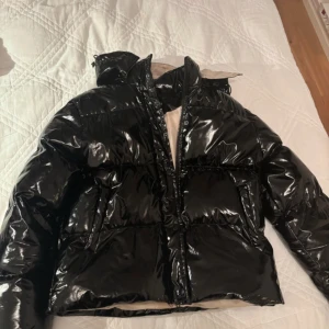 Svart glansig jacka - Säljer en svart glansig pufferjacka med huva och dragkedja. Jackan har en beige insida och en rund logotyp på ärmen. Perfekt för kyliga dagar när du vill hålla stilen. Kan sänka prisen ge bud så kan vi se! 🖤