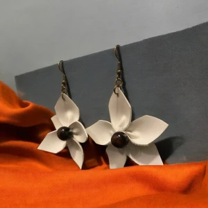 Vita blommiga örhängen - Unika vita örhängen formade som blommor av konstläder, med en mörk träpärla i mitten. Perfekta för att ge en elegant touch till din outfit. De hänger från en enkel krok och är lätta att bära.