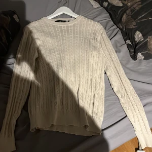 Beige stickad tröja från My Wear - Säljer en stilren beige stickad tröja från My Wear. Tröjan har ett klassiskt flätmönster och långa ärmar. Perfekt för kyligare dagar.