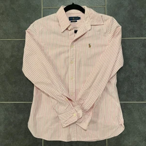 Rosa och vit randig skjorta från Ralph Lauren - Snygg rosa och vit randig skjorta från Ralph Lauren i storlek M. Skjortan har långa ärmar och är broderad med den klassiska logotypen på bröstet. Perfekt för en stilren look!