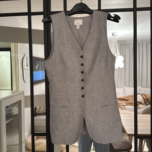 Grå väst med knappar  - Storlek 36 men upplever den liten. Köpt förra juni och använt en enda gång. Snygg grå väst med knappar framtill och två fickor. Perfekt för en stilren look. Västen har en v-ringad design och är tillverkad i ett lätt material.