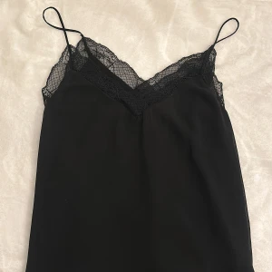 Svart linne med spetsdetaljer - Snygg svart linne med tunna axelband och vacker spetsdetalj vid halsringningen. Perfekt för en stilren och elegant look.
