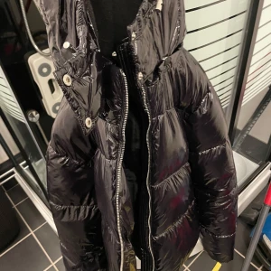 Svart glansig dunjacka - Säljer en svart glansig dunjacka med dragkedja och knappar. perfekt för kyliga dagar. Den är både stilren och funktionell, vilket gör den till ett måste i garderoben. Hör av dig vid intresse! 🖤