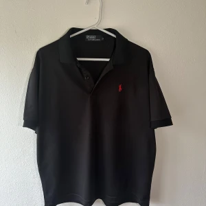 Svart pikétröja från Ralph Lauren - Snygg svart pikétröja från Ralph Lauren med klassisk krage och knappar. Den har en liten röd broderad logotyp på bröstet. Perfekt för en stilren look.