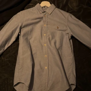 Ljusblå skjorta från Ralph Lauren - Snygg ljusblå skjorta från Ralph Lauren med klassisk krage och broderad logga på bröstet. Skjortan har långa ärmar och knappar framtill. Perfekt för en stilren look.