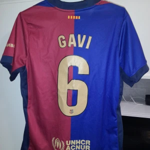 Gavi FC Barcelona fotbollströja - Säljer en blåröd FC Barcelona fotbollströja från Nike med Gavi och nummer 6 på ryggen. Tröjan har klubbens emblem och sponsorlogga på framsidan. Kortärmad design med klassiska färger och detaljer.