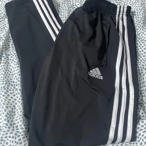 Svarta träningsbyxor från Adidas med Real Madrid-logga - Snygga svarta träningsbyxor från Adidas med de klassiska tre vita ränderna längs sidorna. Byxorna har en broderad Real Madrid-logga på ena benet och elastisk midja för extra komfort.