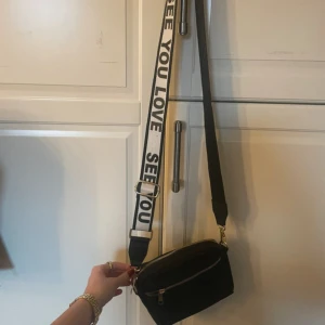 Svart axelväska med text - Snygg svart axelväska med justerbar axelrem som har texten 'SEE YOU LOVE'. Väskan har en dragkedja och är perfekt för att bära med sig det nödvändigaste. Enkel och stilren design som passar till många tillfällen.