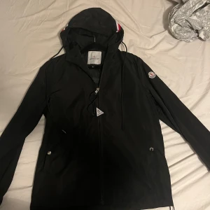 Moncler svart windbreaker storlek S - Säljer en stilren svart vindjacka från Moncler med huva och dragkedja. Jackan har en diskret logga på ärmen och dragsko vid huvan. Perfekt för blåsiga dagar.