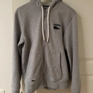 Grå hoodie från Lacoste - Säljer en stilren grå hoodie från Lacoste med dragkedja och justerbar huva. Tröjan har två praktiska fickor framtill och den ikoniska krokodilloggan på bröstet. Perfekt för en avslappnad look.