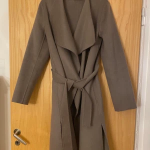 Brun kappa från Wera - Snygg brun kappa från Wera med knytband i midjan. Den har en elegant omlott-design och långa ärmar. Perfekt för kyligare dagar och ger en stilren look. 🧥