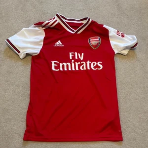 Röd Arsenal fotbollströja från Adidas - Säljer en röd Arsenal fotbollströja från Adidas med vit och svart detaljering. Tröjan har korta ärmar och klubbens emblem på bröstet. Perfekt för alla Arsenal-fans! ⚽