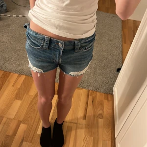 Blå jeansshorts med spetsdetaljer från H&M - Snygga blå jeansshorts från H&M med fransiga kanter och spetsdetaljer. Perfekta för en avslappnad sommarlook. De har en klassisk femficksdesign och knappgylf. Passar Xs