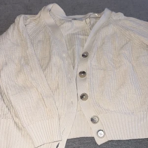 Vit kofta med knappar - Säljer en stilren vit kofta med knappar framtill. Koftan har långa ärmar och ribbade detaljer vid ärmslut och nederkant. Perfekt för lager-på-lager under kyligare dagar.
