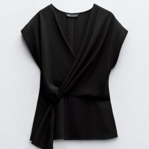 Svart omlottopp från Zara - Snygg svart omlottopp från Zara med en elegant v-ringning och knytning i sidan. Perfekt för en stilren look. Toppen har korta ärmar och är tillverkad i ett mjukt material.