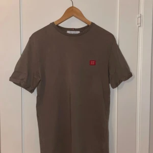 Brun t-shirt från Les Deux - Snygg brun t-shirt från Les Deux med en liten röd broderad detalj på bröstet. Perfekt för en stilren och avslappnad look. Tillverkad i mjukt material för extra komfort.