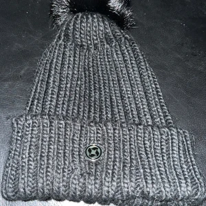 Svart stickad mössa med pälsboll från Colmar - Snygg svart stickad mössa från Colmar med en fluffig pälsboll på toppen. Mössan har en ribbad design och en liten metallknapp framtill. Perfekt för att hålla värmen under kalla dagar.