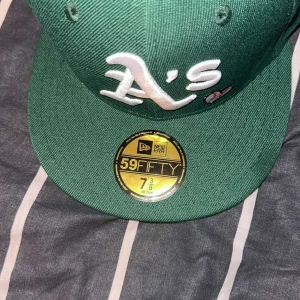 Grön keps från New Era - Snygg grön keps från New Era med vit broderad logotyp på framsidan. Kepsen är en 59FIFTY-modell med en platt skärm och klistermärke kvar på skärmen. Perfekt för att ge din outfit en sportig touch.