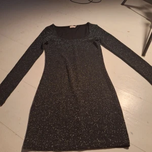Svart glittrig klänning från Nelly - Snygg svart glittrig klänning från Nelly med långärmad design. Perfekt för en kväll ute med vänner. Klänningen har en figurnära passform och en rund halsringning.
