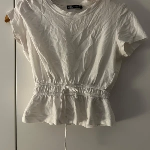 Vit kortärmad topp från Zara - Säljer en söt vit topp från Zara med korta ärmar och en elastisk midja med knytband. Toppen har en lätt volangdetalj vid nederkanten som ger en feminin touch.