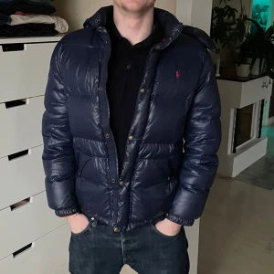 Mörkblå dunjacka från Ralph Lauren - Säljer en snygg mörkblå dunjacka från Ralph Lauren. Jackan har en huva och stängs med både dragkedja och knappar. Perfekt för kyliga dagar med sin quiltade design och långa ärmar. En klassisk och stilren jacka för alla tillfällen. Storleken är S (XL barn)😊