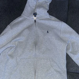 Grå hoodie från Polo Ralph Lauren - Säljer en stilren grå hoodie från Polo Ralph Lauren. Den har en dragkedja framtill och en klassisk logga på bröstet. Perfekt för en avslappnad look. Barnstorlek XL. Köpt för 1200