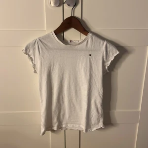 Vit t-shirt från Tommy Hilfiger - Säljer en klassisk vit t-shirt från Tommy Hilfiger. Den har korta ärmar med en subtil volangdetalj och en liten logotyp på bröstet. Perfekt för en stilren och avslappnad look.