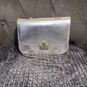 Silverfärgad axelväska med kedja - Snygg silverfärgad axelväska med en glittrig låsdetalj och kedjerem. Väskan har en elegant design och är perfekt för att ge en touch av glamour till din outfit. Insidan är fodrad och har en liten ficka för extra förvaring.