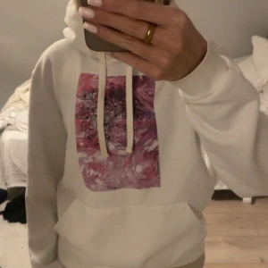 Vit hoodie från The Cool Elephant - Säljer en vit hoodie från The Cool Elephant med en unik konstnärlig design i rosa och lila på framsidan. (flower girl) Hoodien har en stor ficka framtill och justerbar huva med snören. 