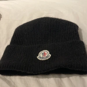 Svart stickad mössa från Moncler - Snygg svart stickad mössa från Moncler med broderad logotyp framtill. Perfekt för att hålla värmen under kyliga dagar. Klassisk design som passar till det mesta.