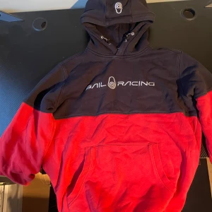 Sail racing hoodie - Säljer en snygg röd och svart hoodie från Sail Racing. Tröjan har en stor logga på bröstet och en praktisk känguruficka framtill. Perfekt för kyliga dagar med sin mjuka och bekväma passform. Perfekt skick och är inte använd