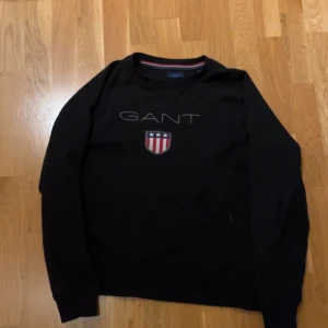Svart sweatshirt från GANT - Säljer en svart långärmad tröja från GANT med ett tryckt emblem på bröstet. Tröjan har en rund halsringning och är perfekt för en casual look. Den har en liten reva under ryggen.                               Kom privat och kan sänka vid snabb affär.                TAR EMOT BYTEN