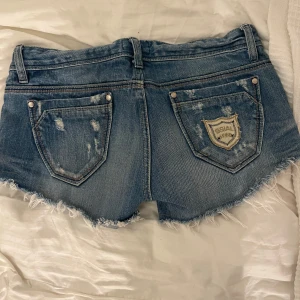 Blå jeansshorts från Ssiajean - Snygga blå jeansshorts från Ssiajean med slitna detaljer och fransig kant. Shortsen har en låg midja och klassiska femficksdesign. Perfekta inför sommaren.💕