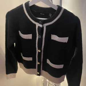 Svart kofta från Vero Moda - Säljer en snygg svart kofta från Vero Moda med vita detaljer och knappar framtill. Använd max två gånger och i jättebra skick. Säljer bara för att jag inte har använt den. Nypris 600kr